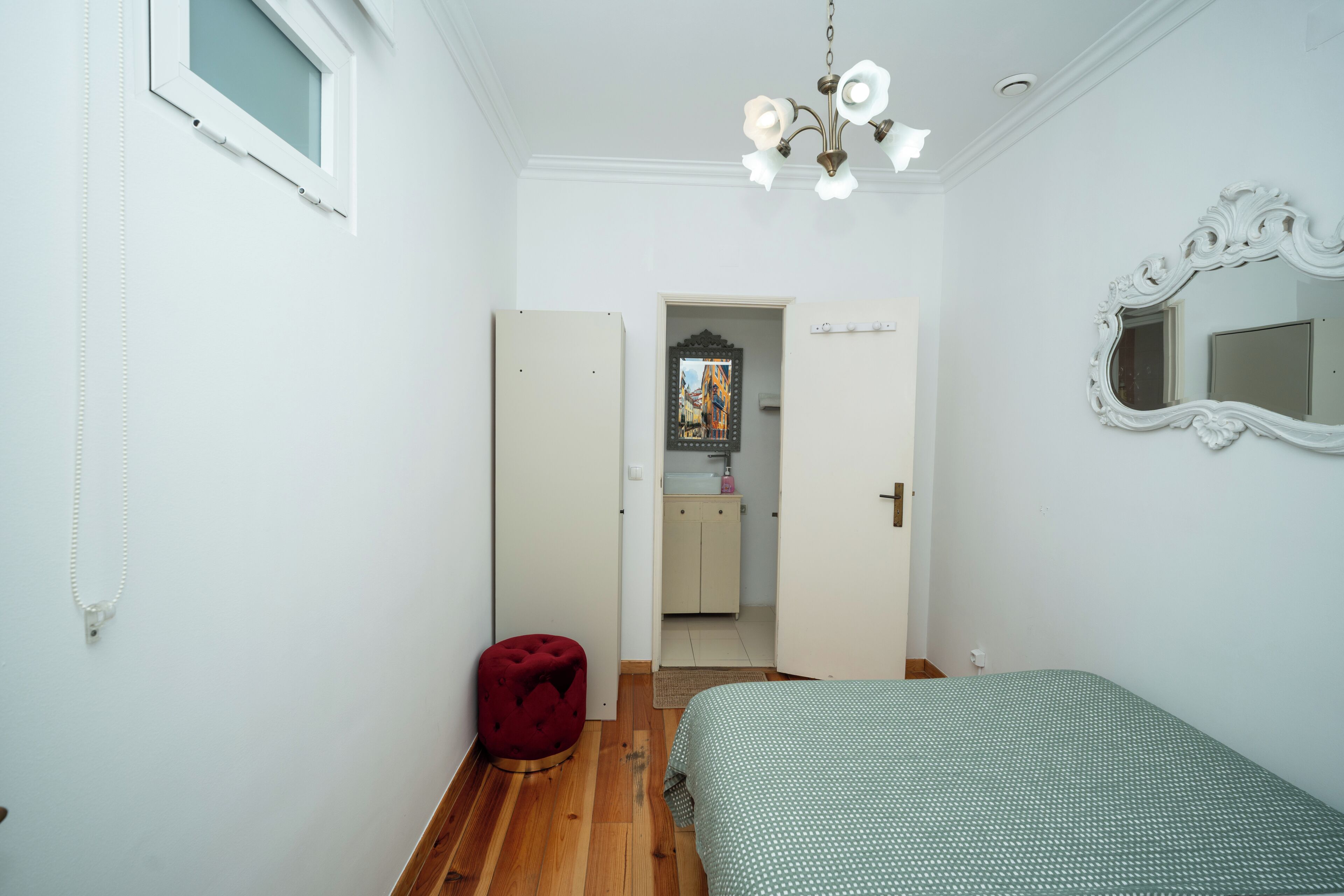 Ein Schlafzimmer mit einem Bett, einem roten Hocker, einem weißen Spiegel und einer Tür zu einem Badezimmer.