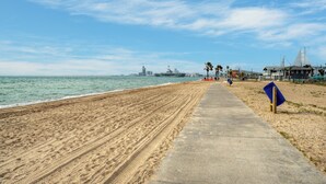 Ein Sandstrand mit einer Betonpromenade, einem blauen Drachen und einem Schiff in der Ferne.