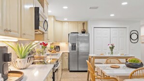 Una cocina moderna con armarios de madera clara, un refrigerador de acero inoxidable y un comedor con sillas de madera y una mesa blanca.