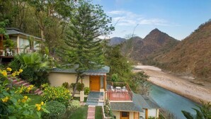 A bungalow with a balcony overlooking a river, surrounded by lush greenery and hills. - Raga on the Ganges (Rishikesh)