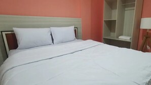 Egy rendezett ágy fehér ágyneművel, egy őszibarack színű falú szobában, mellette egy fa éjjeliszekrényen egy lámpával.