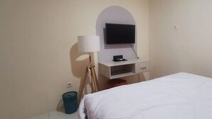 Egy szállodai szoba, amelyben található egy ágy, egy fehér konzolra szerelt televízió, egy lámpa és egy szemeteskosár.