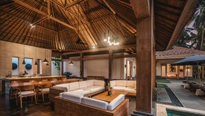 A spacious indoor area with a wooden ceiling, a seating area with a sofa and chairs, a kitchen area with a dining table, and a pool area with lounge chairs. - Villa Jepun (Ubud)