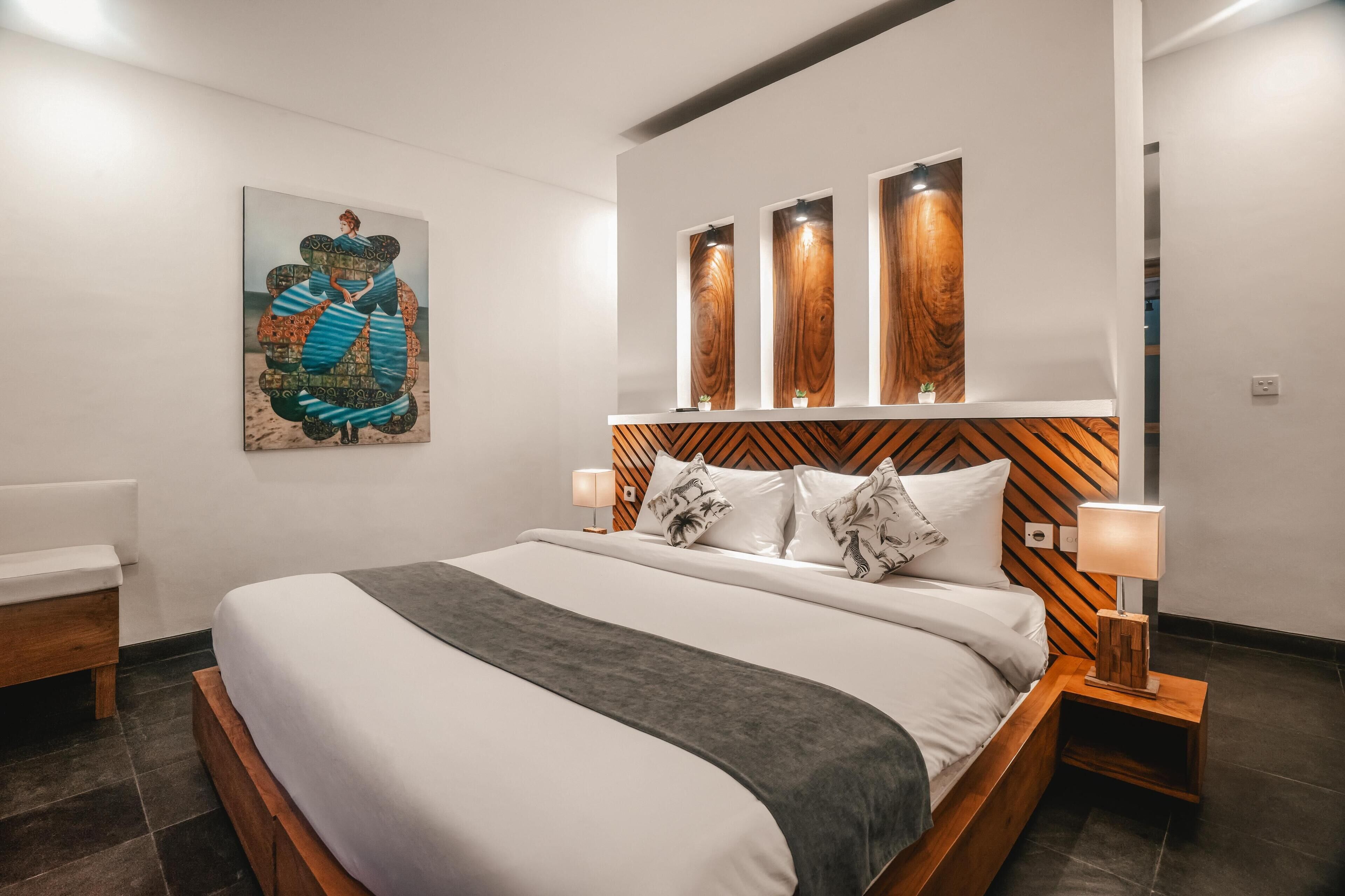 Una habitación de hotel moderna con una cama grande, un cabecero de madera, un cuadro en la pared y una mesita de noche con una lámpara.
