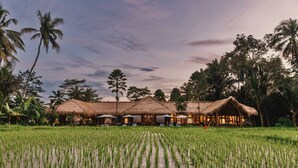 A row of thatched-roof huts with outdoor seating, surrounded by a lush green field. - Villa Jepun (Ubud)