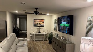 A modern living room with a white leather couch, a TV mounted on the wall, a ceiling fan, and a dining area with a white console table and a plant.