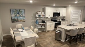 A modern kitchen with white cabinets, a central island with bar stools, a dining area with a table and chairs, and a painting of a beach scene on the wall.