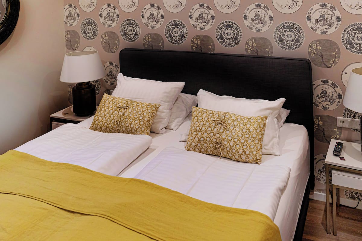 Ein Doppelbett mit weißer Bettwäsche und zwei Zierkissen, ein Nachttisch mit Lampe sowie eine Wand mit ornamentalem Wandpapier.