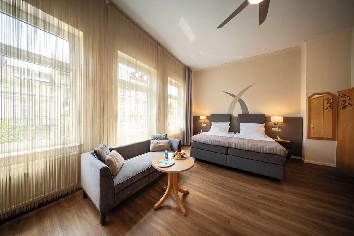 Ein modernes Hotelzimmer mit Sofa, Bett, einem kleinen Tisch mit Früchten und Blick auf die Stadt durch große Fenster.