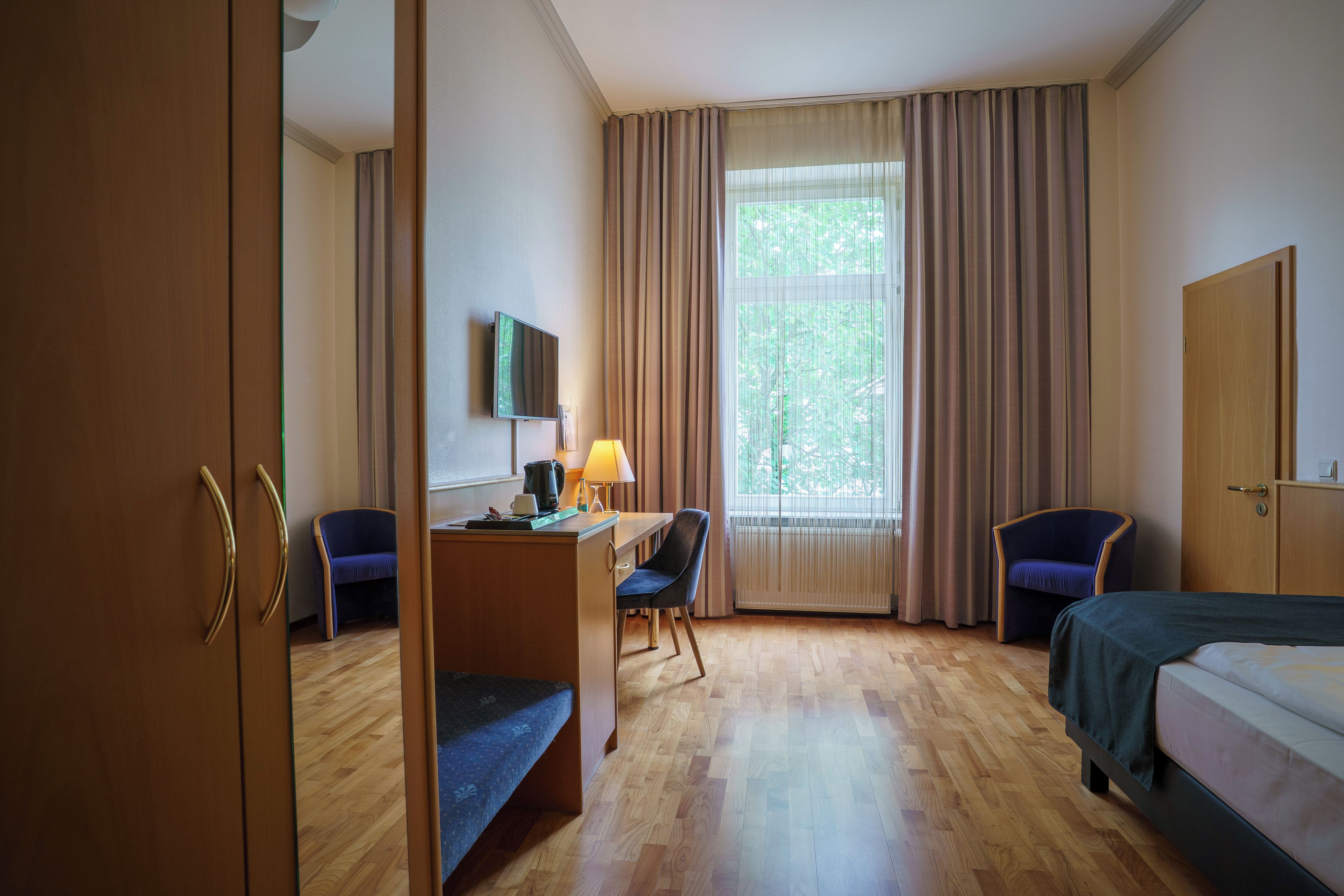 Een hotelkamer met een houten vloer, een bureau met stoel, een bed, een televisie aan de muur en een raam met gordijnen.