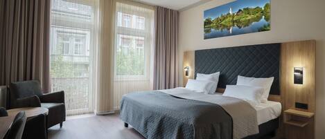 A hotel room with a large bed, a desk, and a chair. There is a painting on the wall and a window with curtains.