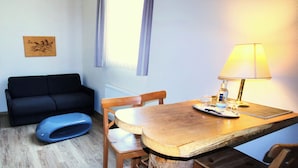 Una habitación pequeña con una mesa de madera, una lámpara, una silla azul, un sofá negro y un radiador.
