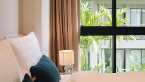 A hotel room with a bed, a lamp, and a view of a building with greenery outside the window. - Solara Stays Phuket (Choeng Thale)