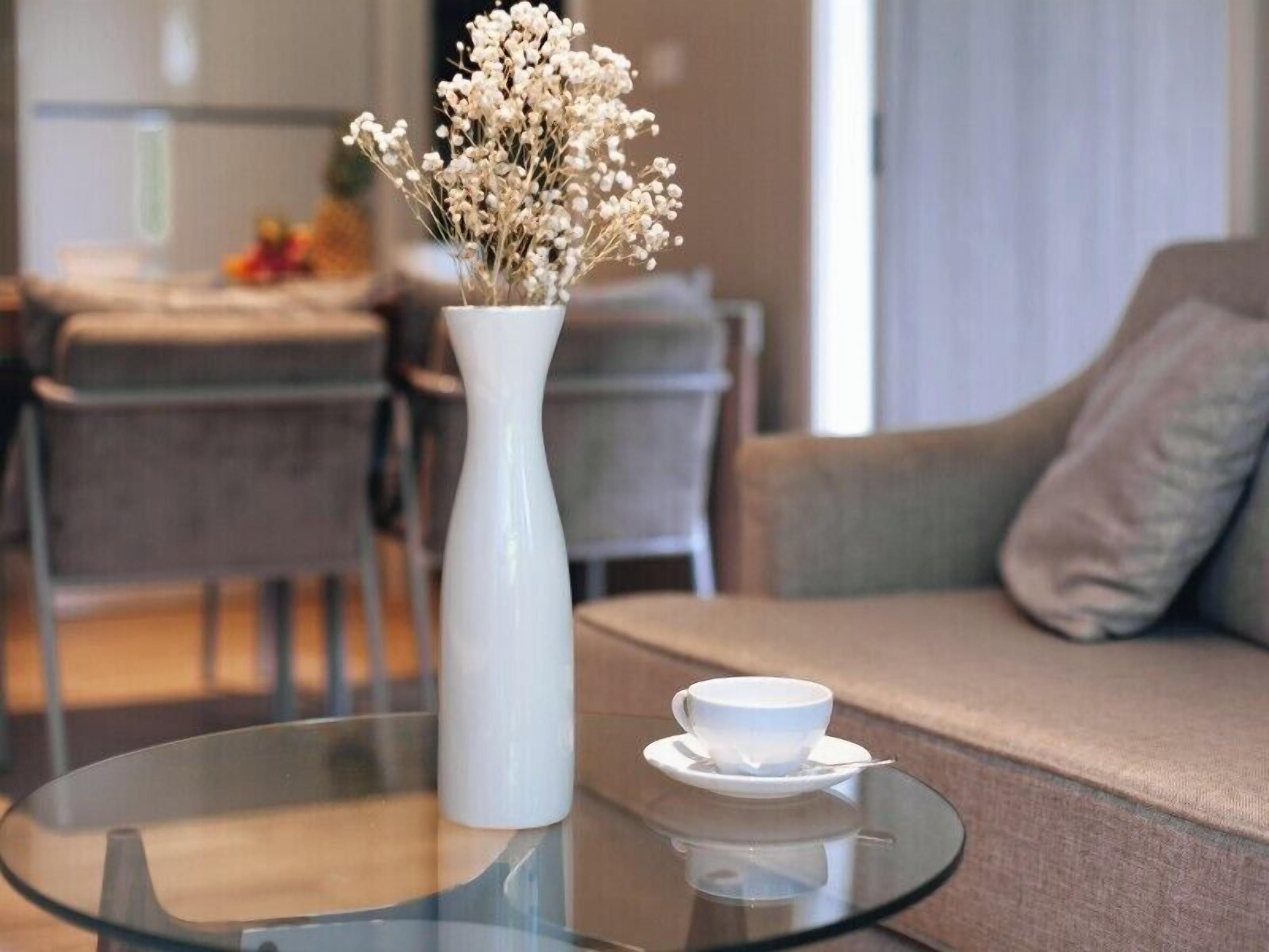 A modern living room with a glass coffee table, a white vase with dried flowers, a white cup and saucer, and a grey sofa.
