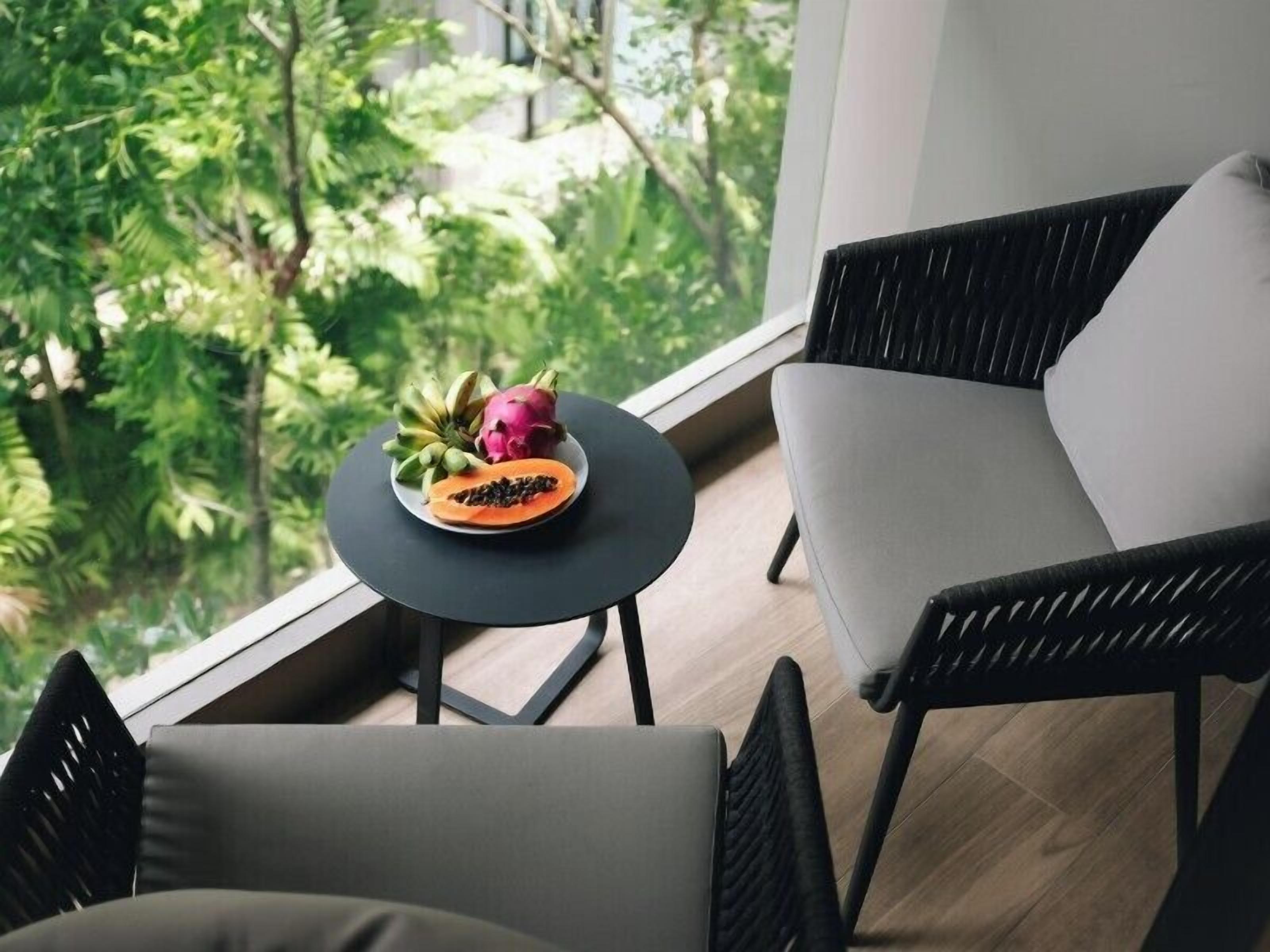 A balcony with a round table holding a fruit bowl, two chairs, and a view of lush greenery.