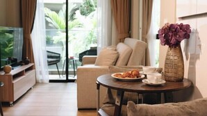 A modern living room with a sofa, a coffee table, and a TV. There is a balcony with plants visible through the glass doors. - Solara Stays Phuket (Choeng Thale)