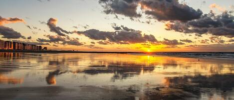 A beach with a sunset, reflecting buildings and clouds in the wet sand.