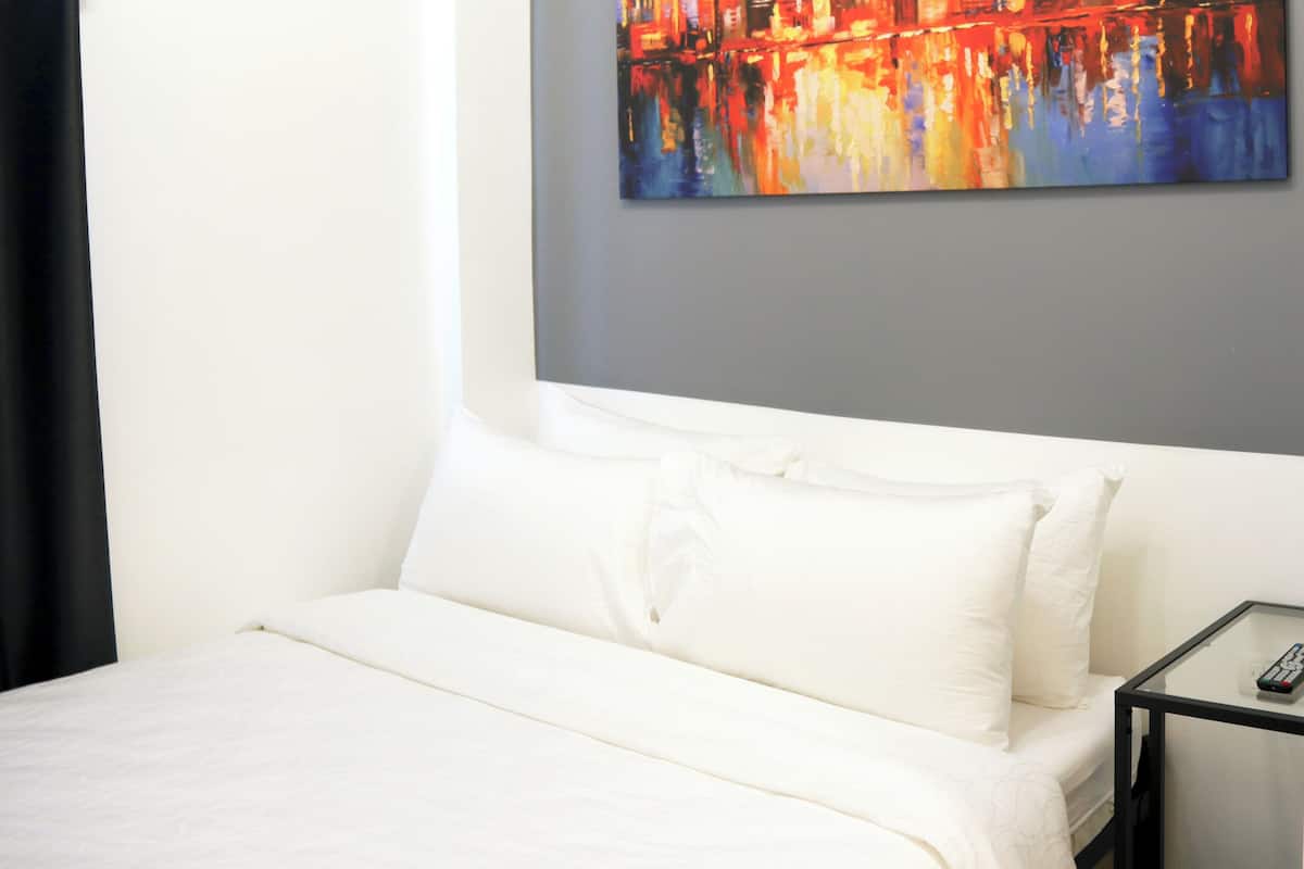 A hotel room with a bed, white bedding, a glass side table with a remote control, and a colorful abstract painting on the wall.