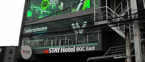 Ein Hotel mit einer großen digitalen Anzeige, die ein Fitnessstudio bewirbt, einem Schild zur Steuerung und dem Namen "STAY Hotel BGC East".