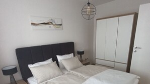 A bedroom with a large bed, a bedside table with a lamp, a wardrobe, and a wall-mounted artwork.