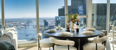 Un coin repas avec une table ronde dressée pour quatre personnes, de grandes fenêtres offrant une vue sur le paysage urbain et un pont, et un panneau de la police apposé sur la fenêtre.