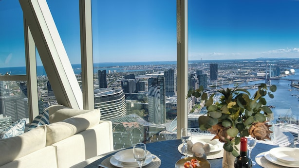 A dining table set with plates, glasses, and a bottle of wine, overlooking a cityscape with modern buildings and a body of water.