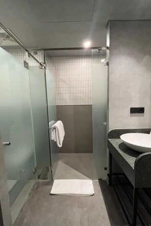 A modern bathroom with a glass shower enclosure, a white towel hanging on the door, and a large sink with a granite countertop. - ATOOI Hotel Alam Sutera (South Tangerang)