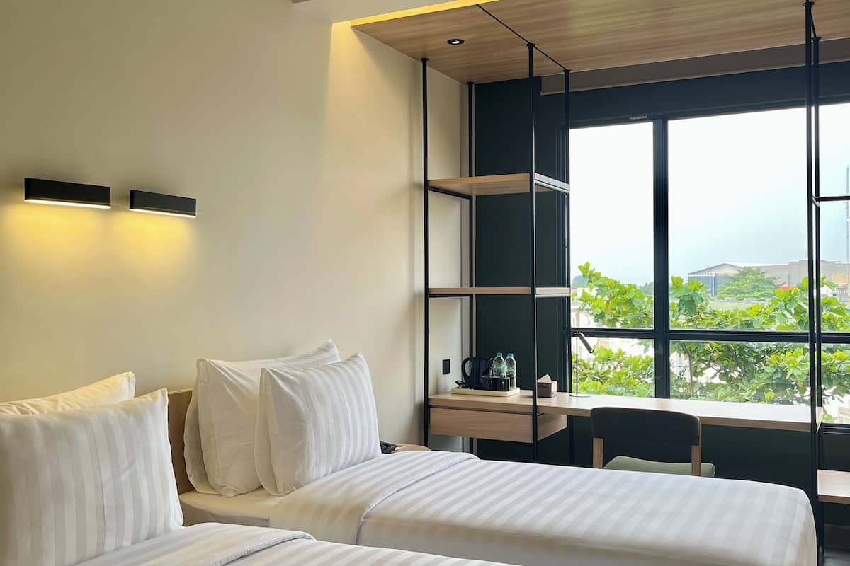 A modern hotel room with two beds, a desk, and a large window offering a view of greenery.