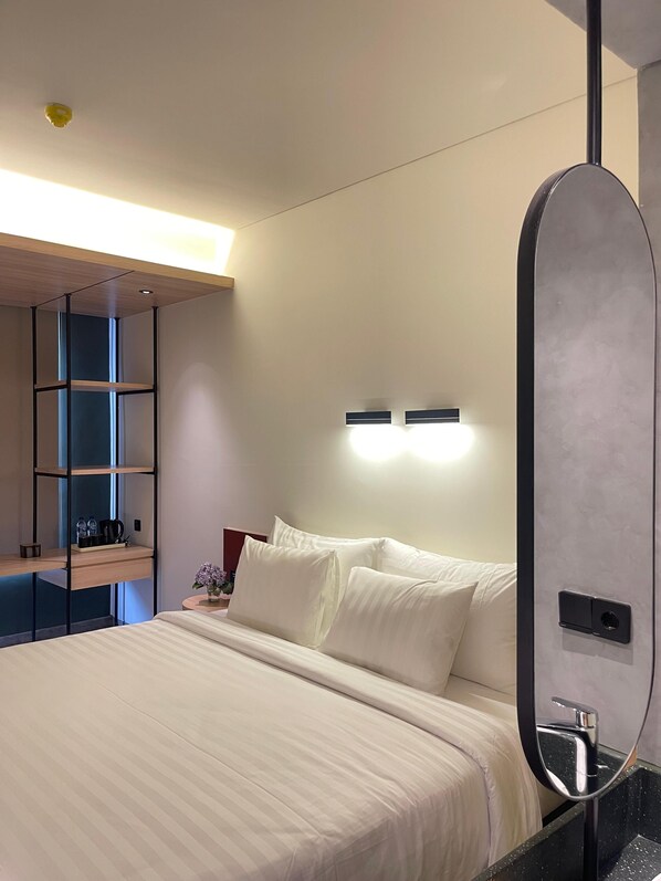 A modern hotel room with a large bed, a built-in shelf unit, a bedside table with a vase of flowers, and a wall-mounted mirror. - ATOOI Hotel Alam Sutera (South Tangerang)