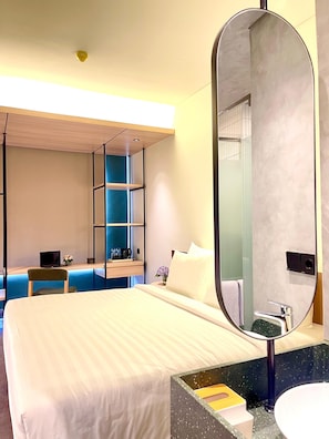 A modern hotel room with a large bed, a desk with a laptop, a blue seating area, and a bathroom with a large mirror and sink. - ATOOI Hotel Alam Sutera (South Tangerang)