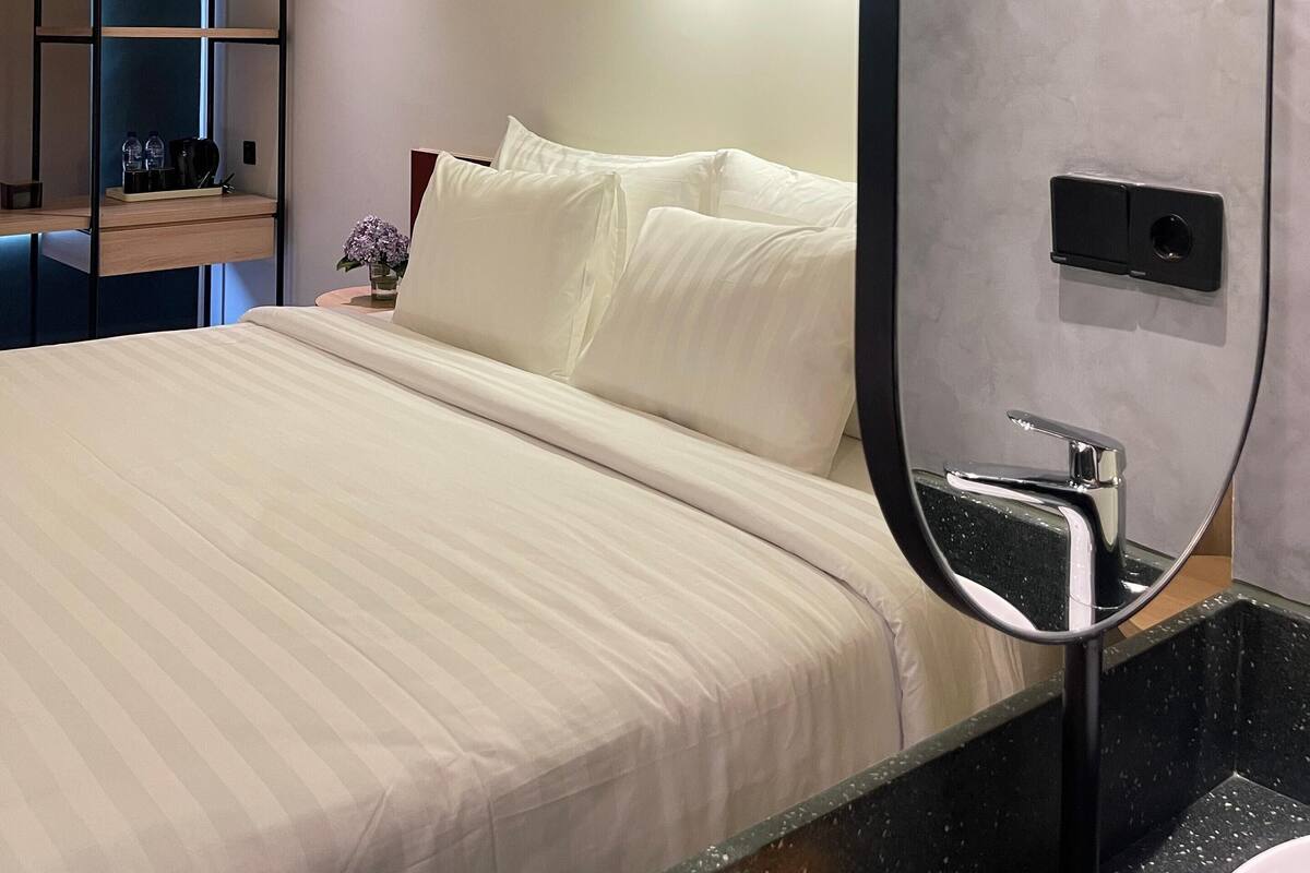 A modern hotel room with a large bed, a built-in shelf unit, a mirror, and a bathroom with a black countertop.