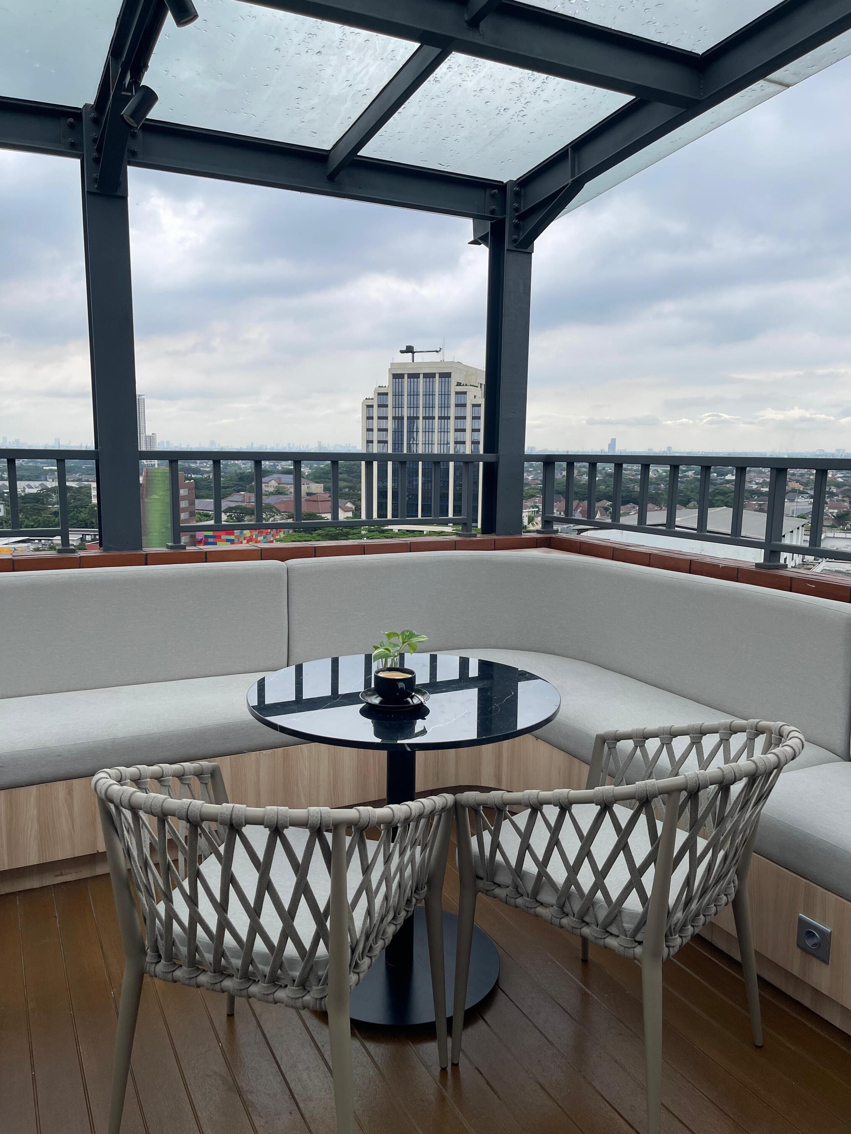 A covered outdoor seating area with a round table, cushioned chairs, and a view of a cityscape.