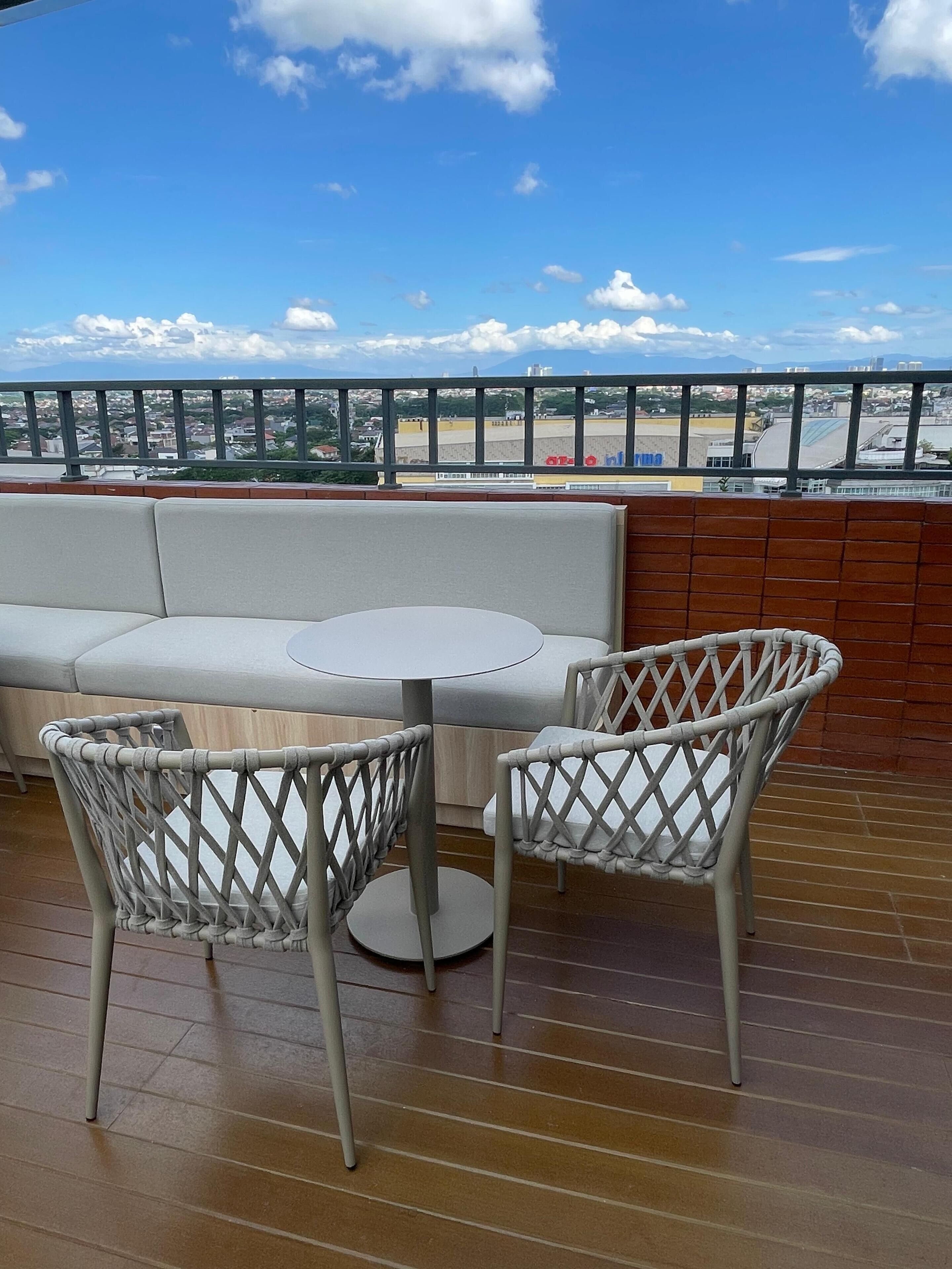 Ein Balkon mit einer weißen gepolsterten Bank und zwei Korbstühlen mit Blick auf eine Stadtlandschaft und einen Strand.