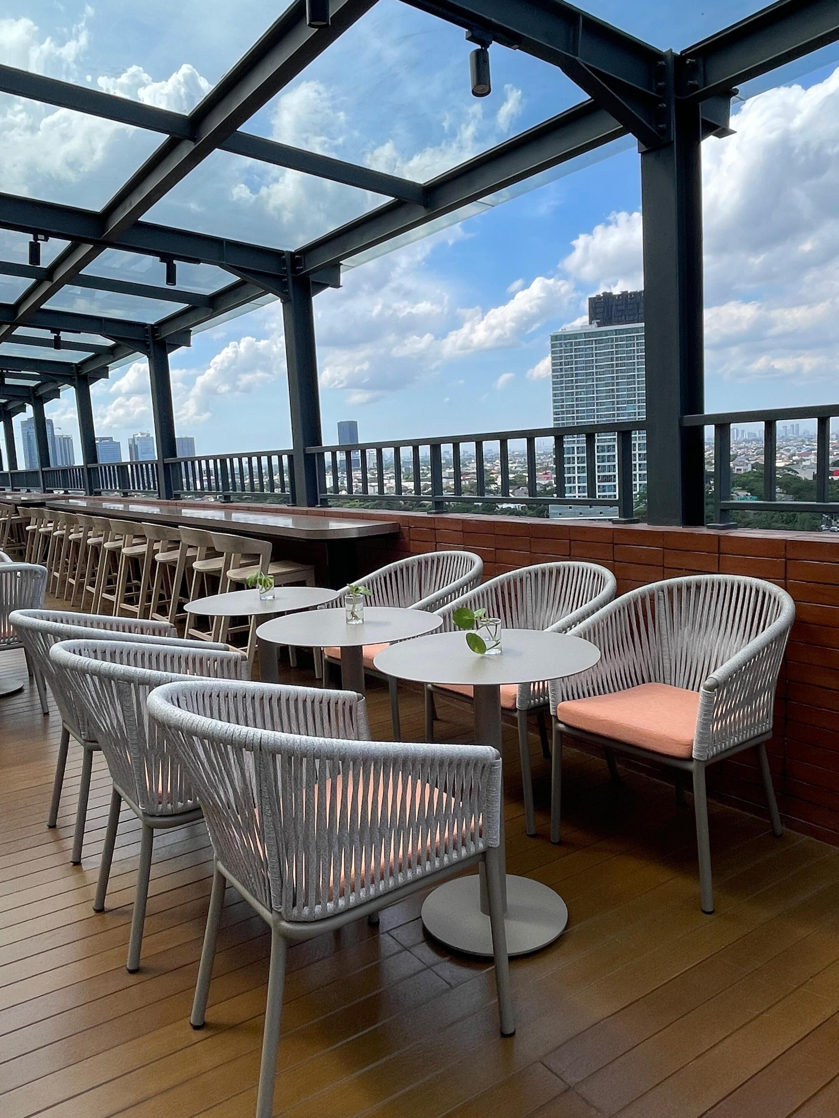 An outdoor seating area with wicker chairs and round tables, overlooking a cityscape with tall buildings.