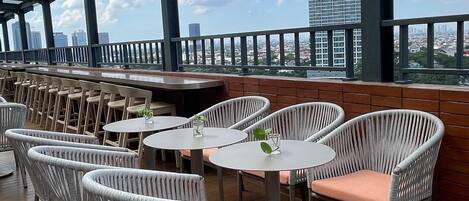 An outdoor seating area with wicker chairs and round tables, overlooking a cityscape with tall buildings.