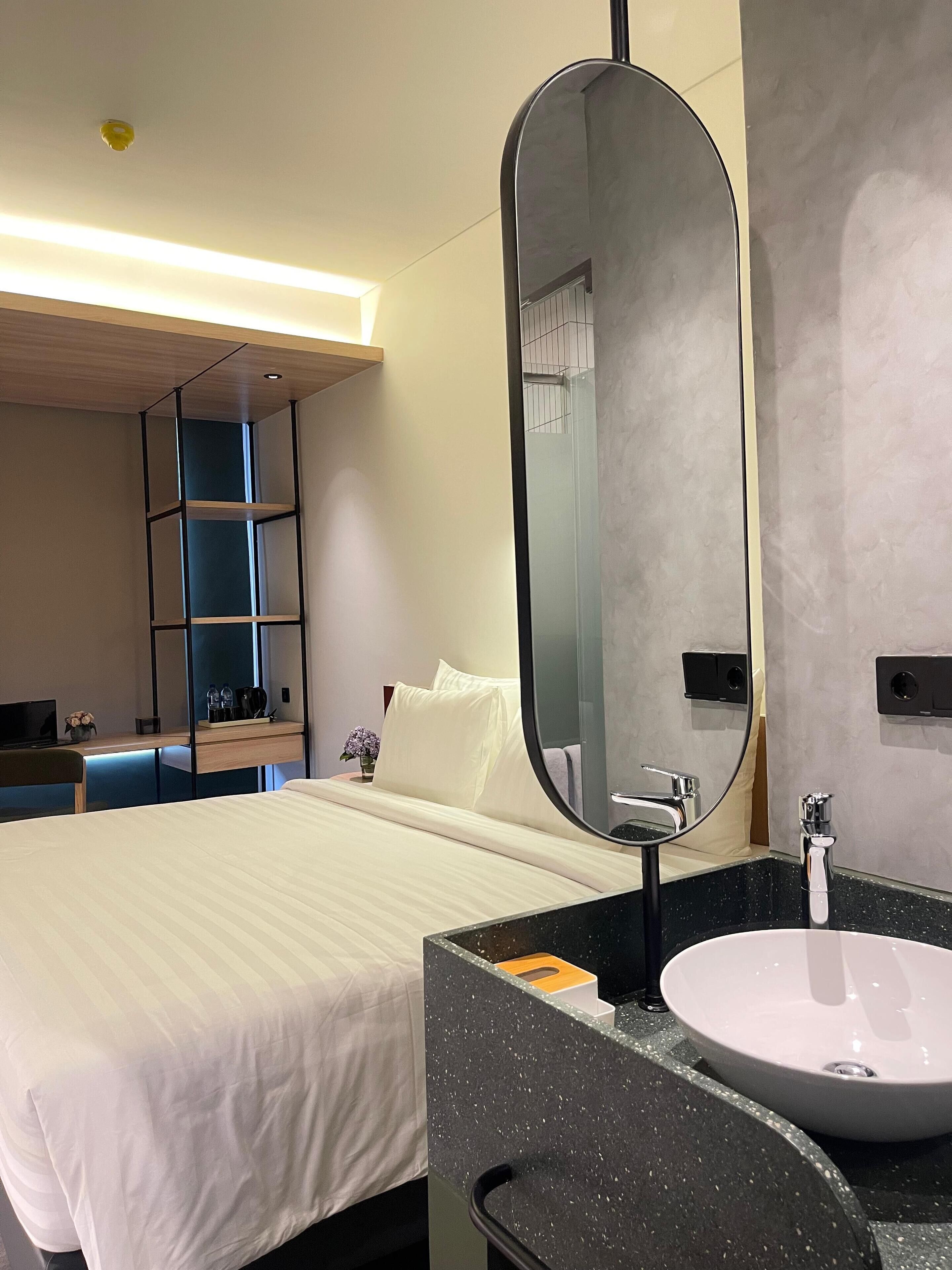 A hotel room with a bed, a round mirror, a black countertop with a sink, and a small desk with a chair.