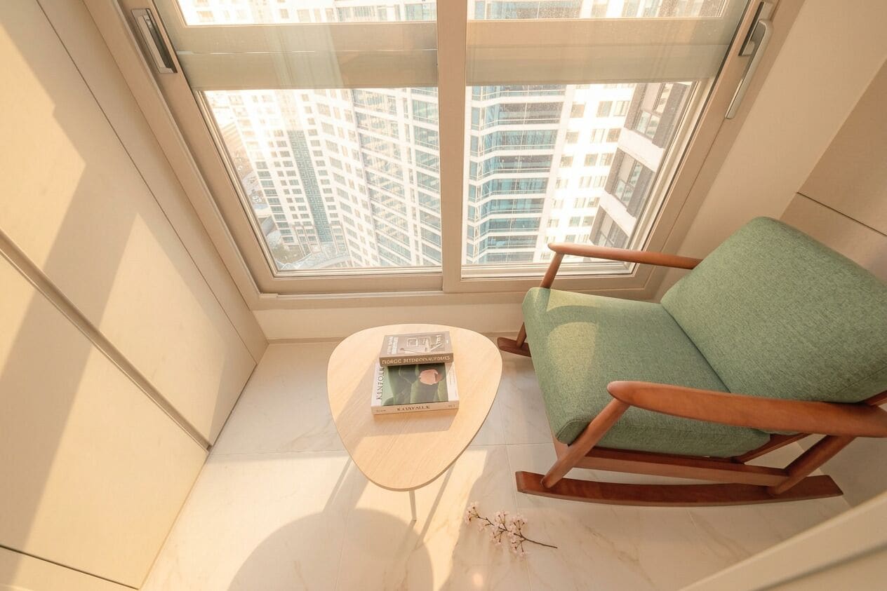 A corner room with a view of high-rise buildings, a rocking chair, a small table with a book, and a vase with flowers.