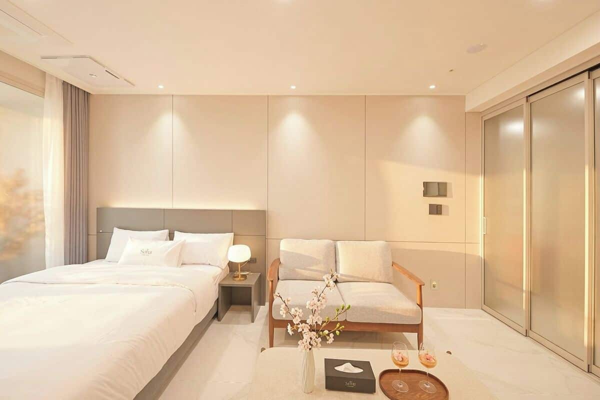 A modern hotel room with a large bed, a sitting area with a chair, a nightstand, a vase with flowers, and a small table with a book and glasses.
