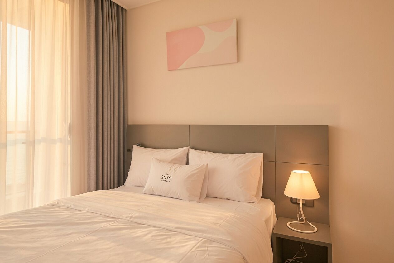 A hotel room with a bed, a nightstand with a lamp, a painting on the wall, and sheer curtains on the window.