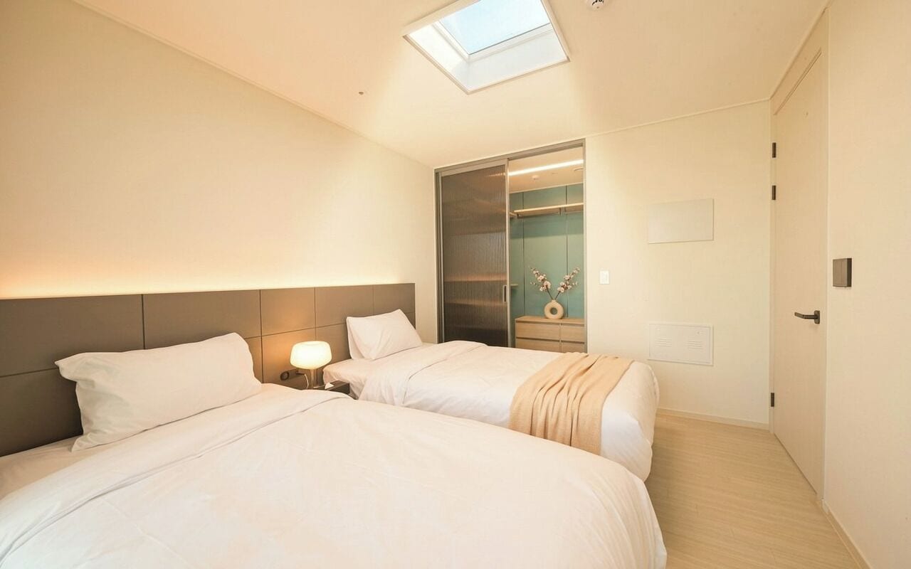 A hotel room with two beds, a skylight, a closet with a mirror, and a bedside table with a lamp.