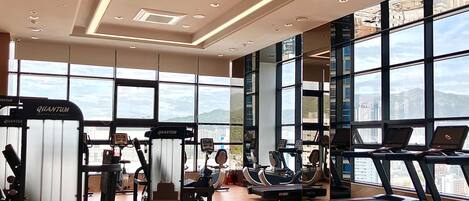 A gym with treadmills, elliptical machines, and weightlifting equipment. Large windows offer views of the cityscape.