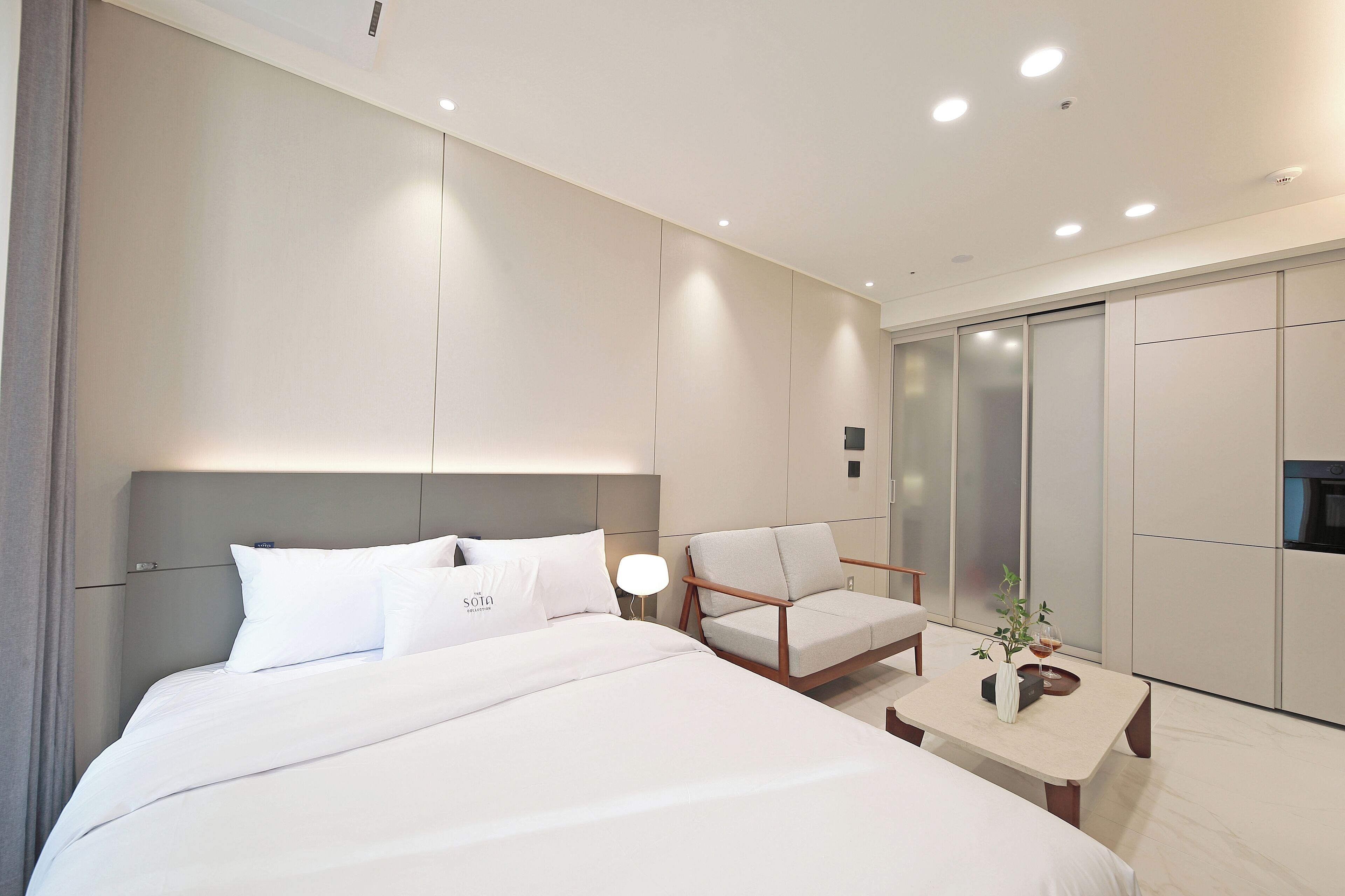 A modern hotel room with a large bed, a seating area with a table, and a sliding door closet.