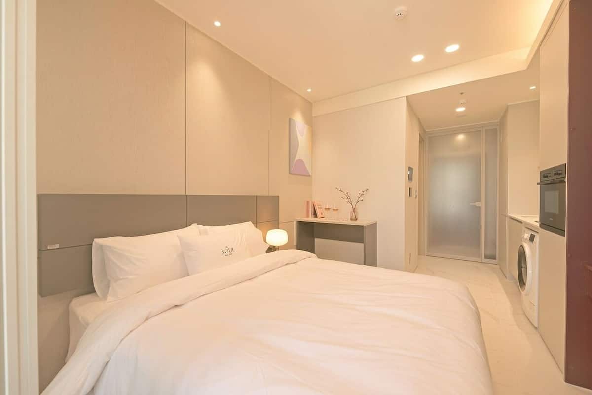 A modern hotel room with a large bed, a bedside table with a lamp, a small shelf with decorative items, and a washing machine in the corner.