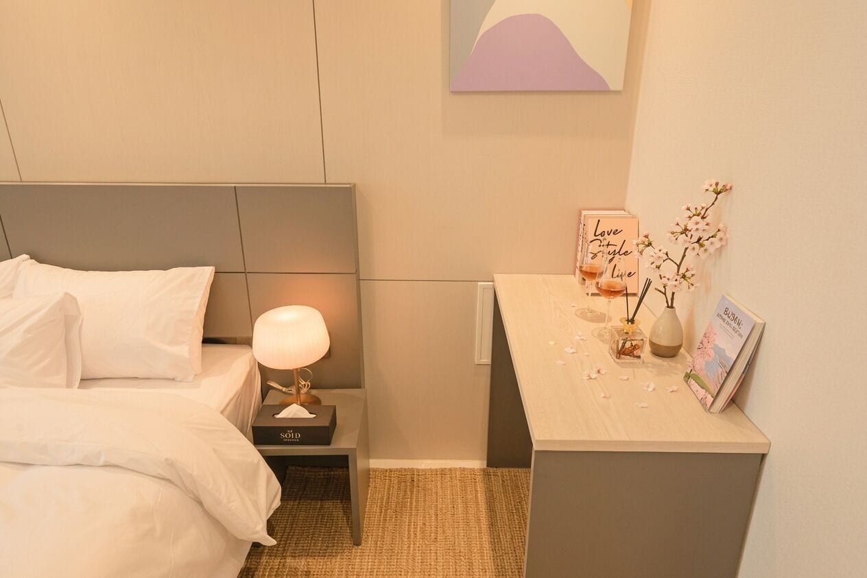 A hotel room with a bed, bedside table with a lamp, a small desk with decorative items, and a wall-mounted artwork.