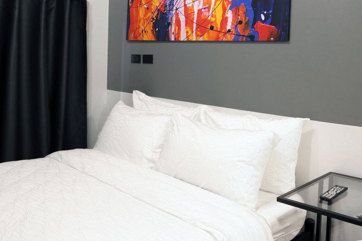 A hotel room with a bed, a glass table, and a colorful abstract painting on the wall.