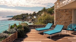 A wooden deck with two lounge chairs and a table, overlooking a coastal area with a stone building and a view of the sea.