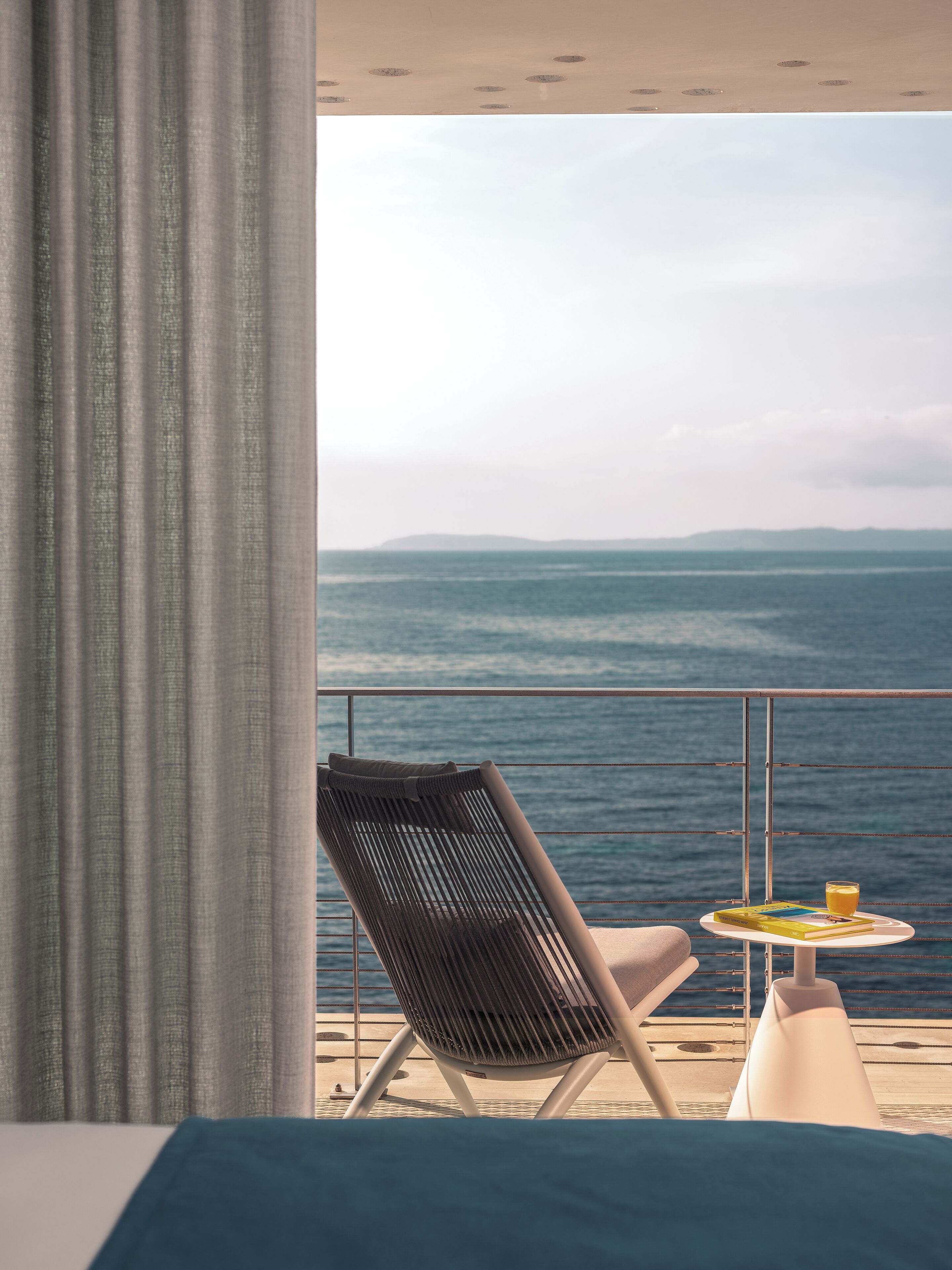 A balcony with a view of the sea, a chair, a small table with a book and a glass of orange juice.