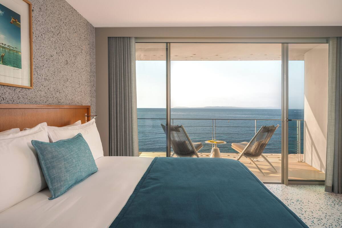 Habitación de hotel con una cama grande, vista al mar y un balcón con sillas.