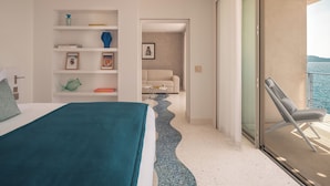 A modern hotel room with a bed, a built-in bookshelf, a small seating area, and a balcony with a view of the sea.