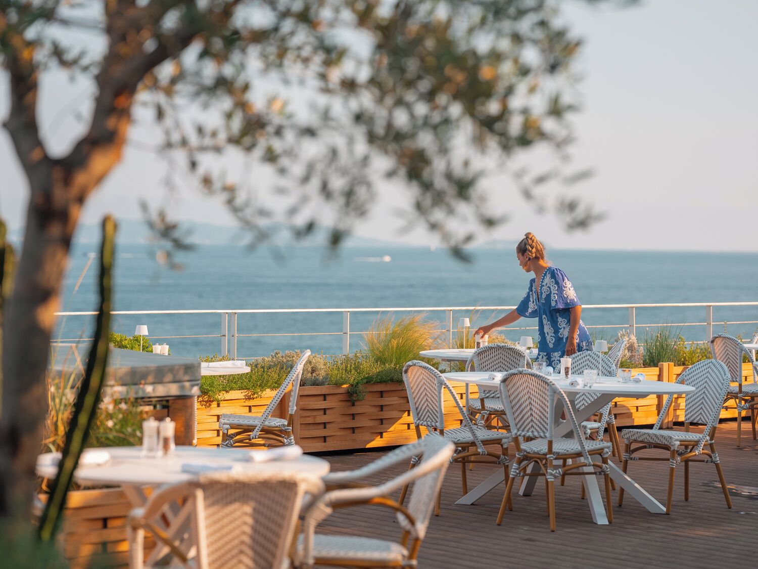 Un restaurant en bord de mer, avec une terrasse, une vue sur l’océan et un serveur qui s’occupe des tables.